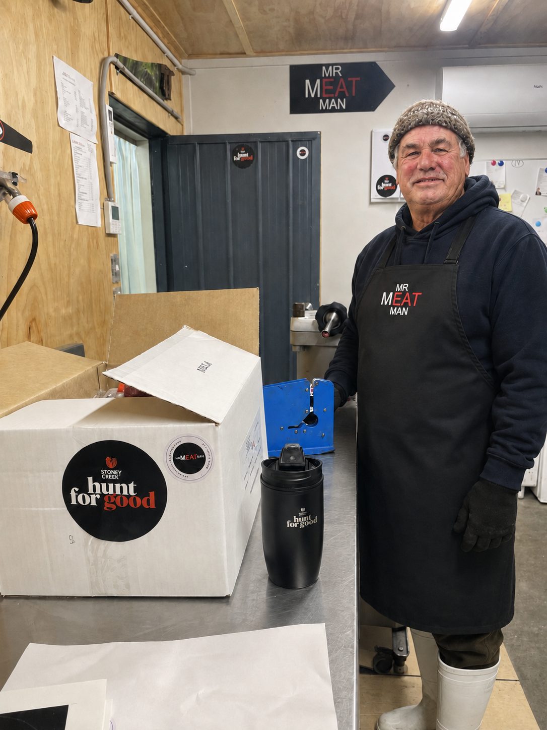 Bruce Richards in his processing room, wearing a Mr Meat Man apron and standing beside a Hunt for Good donation box.